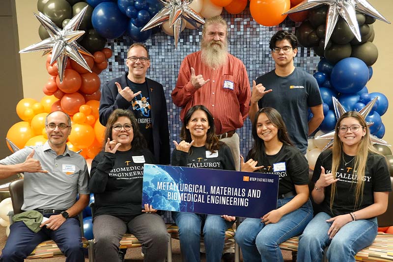 Group of attendees pose together during a UTEP Engineering homecoming gathering, standing in front of a decorative backdrop with orange, blue, black, and silver balloons while holding foam finger props and making hand gesture signs for a celebratory group photo.